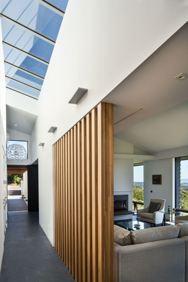 A bright contemporary residential interior showing a long hallway beside a living room. A full-height timber slatted screen forms a warm divider, while a large skylight brings daylight into the circulation space. The living area contains a large neutral sectional sofa, armchair, glass coffee table and fireplace, with expansive windows overlooking a green landscape. Dark flooring contrasts with white walls and timber detailing.