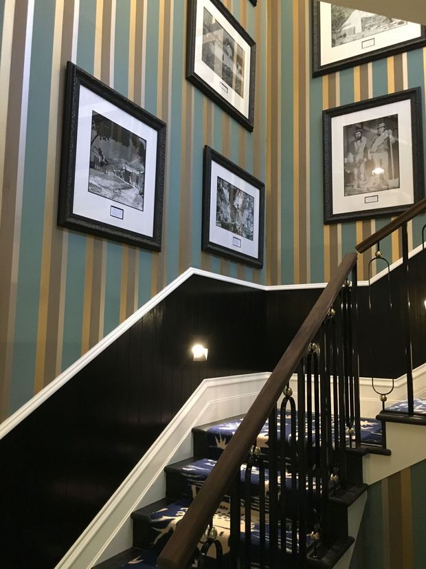 A traditional stairway with black painted wainscoting, white trim, dark timber handrails, and ornate black metal balustrades. The walls feature bold vertical striped wallpaper in muted teal, olive, beige, and cream, with several framed black-and-white photographs displayed along the stairwell. The stairs are carpeted in a dark patterned runner, creating a refined, heritage-inspired interior.