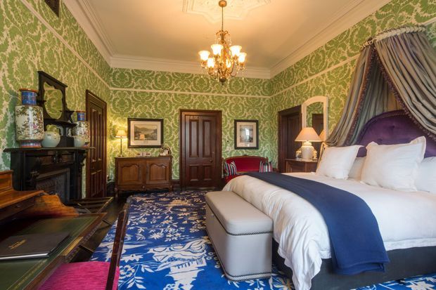 A richly decorated traditional bedroom with ornate green patterned wallpaper, dark timber doors and furniture, a large upholstered bed with a draped canopy, white bedding and a navy bed runner. A blue patterned rug, chandelier, lamps, framed artwork, and decorative shelving create a luxurious, historic interior atmosphere.