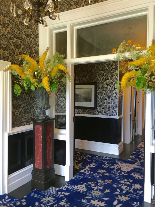 Ornate formal hallway or entry foyer with black-and-white damask wallpaper, black wainscoting, crisp white mouldings, framed artwork, dark flooring, and a richly patterned blue runner. A decorative pedestal with a large floral arrangement and an elaborate crystal chandelier create a dramatic, traditional presentation. Doorways and mirrored glazing extend the corridor into adjoining spaces.