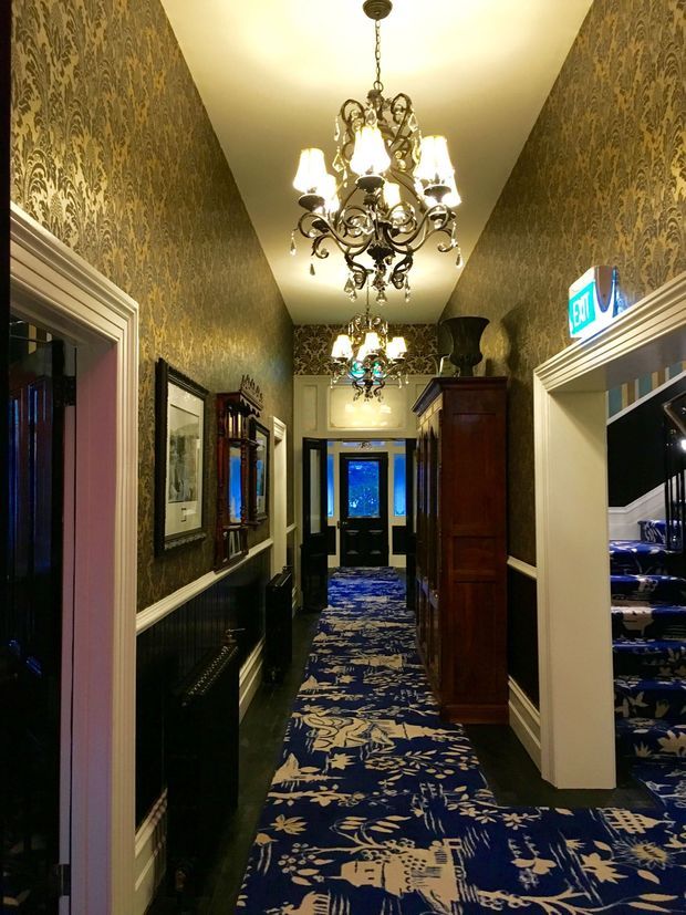 A narrow, richly decorated hallway with dark patterned wallpaper, ornate white trim and wainscoting, multiple elaborate crystal chandeliers, framed artwork, a tall dark wood cabinet, and a bold blue floral-patterned carpet. A staircase is visible through an opening on the right, and glazed entry doors appear at the far end.