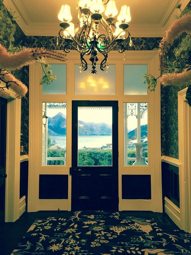 A richly decorated traditional entryway with a dark central glazed door, matching sidelights and frosted transom panels, framed by ornate cream-coloured mouldings and dark patterned wallpaper. A large elaborate chandelier hangs from a coffered ceiling, while a bold floral carpet and decorative wall-mounted greenery add visual detail. Through the glazing, a bright mountain-and-lake landscape is visible.
