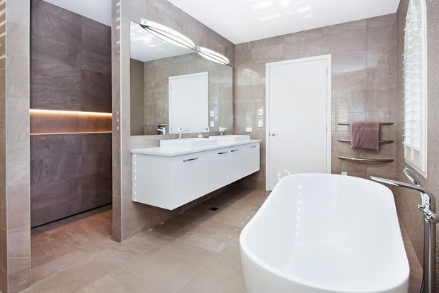 A bright, contemporary bathroom with full-height taupe-brown stone-look wall and floor tiles. The room features a freestanding white bathtub, wall-mounted white vanity with double basins, large mirror, chrome tapware, a glass-enclosed shower with a dark tiled feature wall and recessed warm LED shelf lighting, a white internal door, and a window with white plantation shutters.