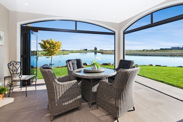 Bright enclosed sunroom or conservatory with expansive arched black-framed glazing overlooking a calm waterfront and lawn. The space contains a round outdoor-style wicker dining set with four armchairs, a small side chair, tiled flooring, recessed ceiling lights, and warm neutral walls. The architecture is defined by curved window heads and generous floor-to-ceiling glazing, creating an airy indoor-outdoor connection.