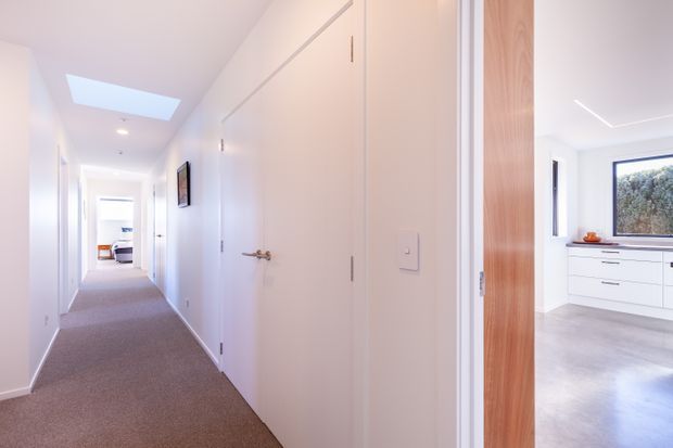 Bright, minimalist residential hallway with white walls, multiple flush interior doors, recessed lighting, a skylight, and soft carpet flooring. An open doorway reveals part of a contemporary white kitchen or utility space with timber detailing, pale flooring, and a large window.