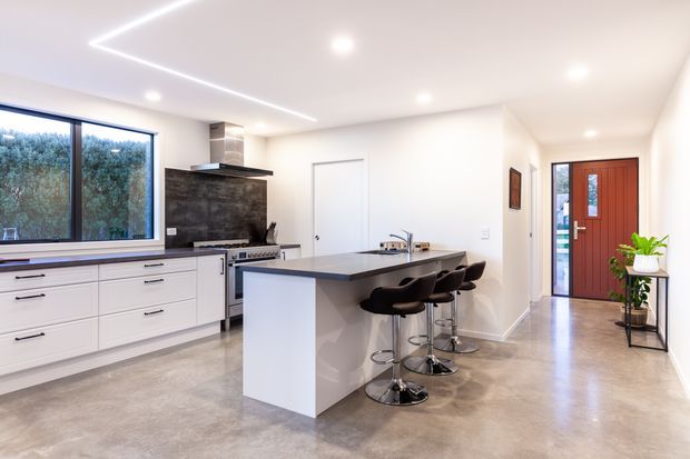 Bright contemporary open-plan kitchen with a large white island, dark benchtop, four bar stools, extensive white drawer cabinetry, and a dark tiled splashback behind the cooktop. The room has polished concrete flooring, recessed ceiling lights, linear LED lighting, large glazing, and a timber front door visible at the end of the space.