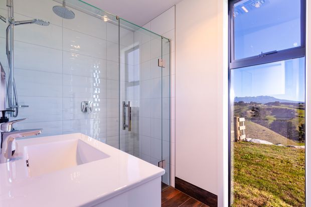 Bright contemporary bathroom with a freestanding white bathtub, wall-mounted chrome fixtures, a glass-enclosed walk-in shower, glossy white wall tiles, and dark wood-look flooring. A large window provides expansive views of the surrounding rural landscape and hills.