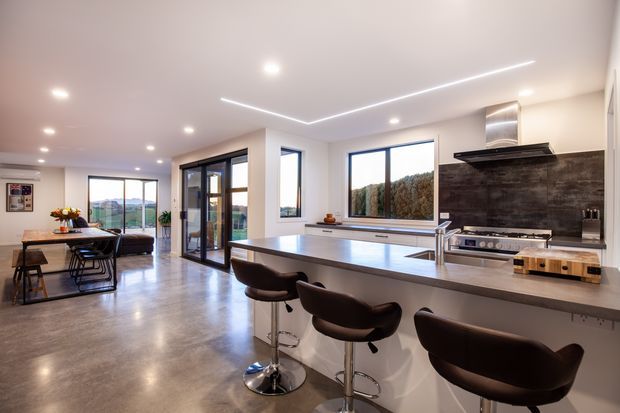 A spacious open-plan contemporary kitchen and dining/living area with a large island, integrated seating, dark cabinetry, stainless-steel appliances, and a dark textured splashback. Polished concrete flooring, white walls, recessed lighting, and linear LED ceiling details create a bright, modern interior. Floor-to-ceiling glazing and sliding doors connect the space to the outdoors, with a dining table and lounge area visible beyond the kitchen.