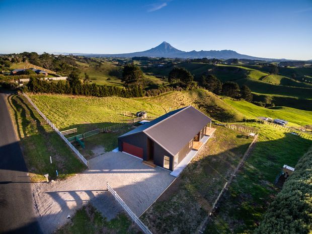 Aerial view of a rural property with a modern dark-clad barn-like outbuilding, gravel forecourt, fenced paddocks and rolling green farmland. A distinctive volcanic mountain rises in the distant landscape under a clear blue sky.