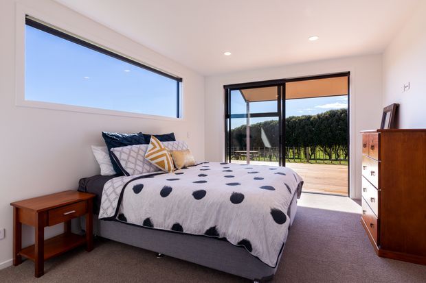Bright contemporary bedroom with a low upholstered bed, patterned bedding, timber bedside table and dresser, pale walls, carpeted floor, and large black-framed windows and sliding glass doors opening to a landscaped outdoor area.