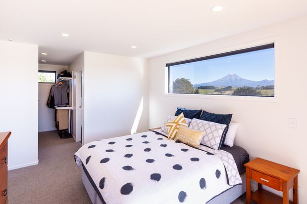 Bright, compact contemporary bedroom with a double bed, patterned navy-and-white bedding, colourful accent cushions, recessed ceiling lights, carpeted flooring, and a built-in open wardrobe area near the entrance. A wide horizontal window frames a clear mountain landscape.