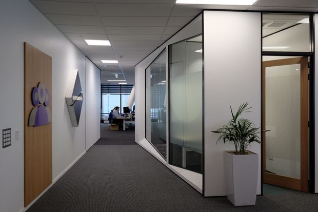 A modern commercial office corridor with dark carpet flooring, white walls, glazed meeting-room partitions, timber doors, recessed fluorescent ceiling lighting, and a potted plant. Workstations are visible at the far end, while geometric wall artwork and signage add subtle visual accents.