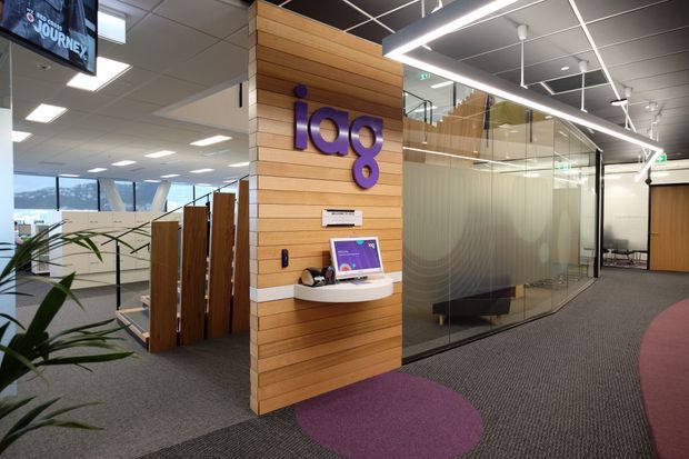 Contemporary commercial office reception or circulation area featuring a timber-lined feature wall with purple branding and a small digital kiosk. Glazed meeting rooms with frosted lower panels, exposed dark ceiling grid, linear LED lighting, carpeted flooring with circular purple accents, and open-plan work areas are visible.