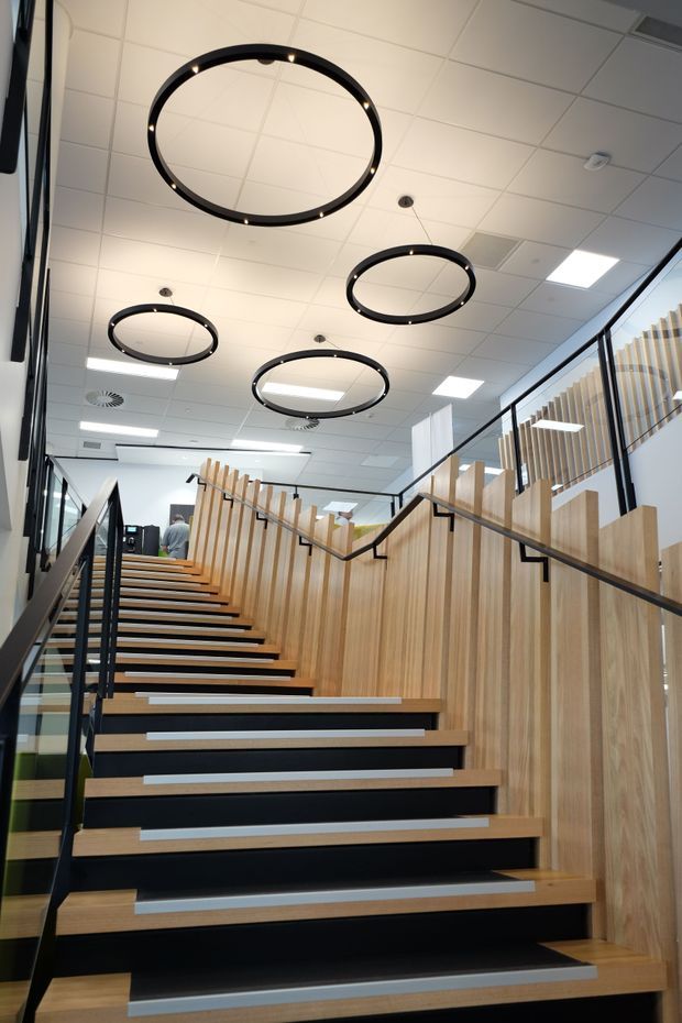 A contemporary commercial interior showing a broad staircase rising through an open office or institutional space. The stair has light timber treads and vertical timber screening, contrasting with black risers, slim black handrails, and glass balustrades. Circular black pendant lights hang beneath a white suspended ceiling with recessed grid panels and fluorescent fixtures. The image emphasizes the stairway, balustrade, timber detailing, and bright modern workplace atmosphere.