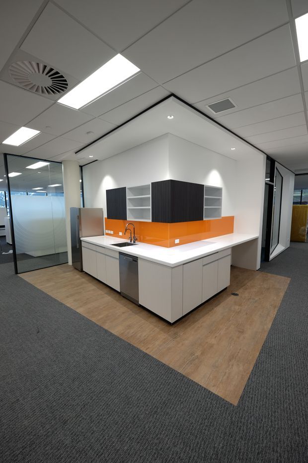 Modern commercial office kitchenette in an open-plan workspace, featuring a central white island with integrated sink and dishwasher, dark wood-grain overhead cabinetry, open shelving, and a vivid orange glass or laminate splashback. The area is surrounded by glass-partitioned offices and finished with white acoustic ceiling tiles, recessed lighting, grey carpet, and a timber-look floor inset.