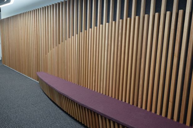 A modern interior corridor or waiting area featuring a long built-in curved bench upholstered in muted purple fabric. The wall behind it is clad with closely spaced vertical timber slats that vary in height to create a stepped, wave-like feature. Dark acoustic backing is visible between the slats, and the floor is covered in dark grey carpet. A recessed linear ceiling light is partially visible.