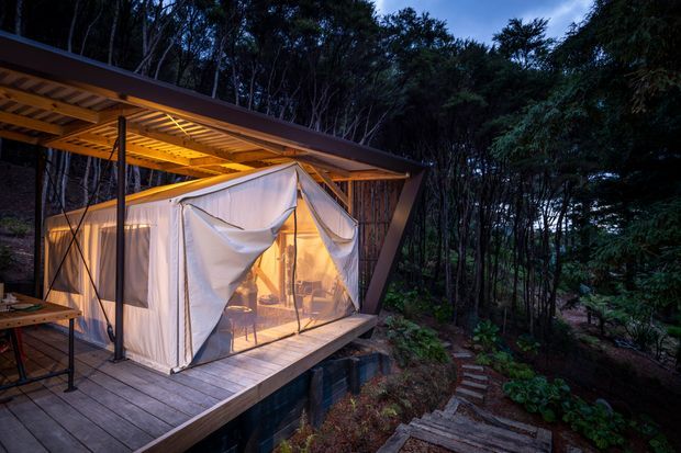 A small elevated tent-like shelter with translucent fabric walls and warm interior lighting, set on a timber deck among dense native woodland. The structure has a broad sloping roof with exposed timber rafters, dark slender supports, and a stepped path leading through the landscaped forest.