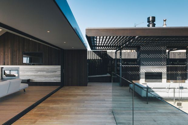 Contemporary indoor-outdoor living space with dark vertical timber wall cladding, warm timber flooring, a low-profile lounge area, and a covered terrace separated by frameless glass balustrading. A timber pergola creates patterned shadows over the outdoor seating and fireplace area, with clear blue sky visible above.