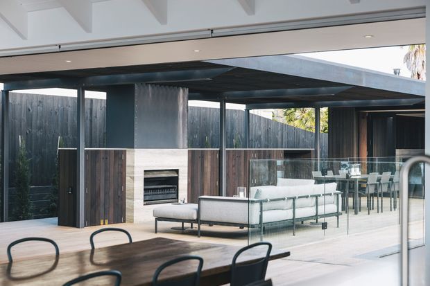 Open-plan indoor-outdoor living and dining area with a contemporary pavilion-like design. The space features a dark timber-clad fireplace wall, black steel structural posts, extensive glazing, a linear outdoor fireplace, light timber flooring, and a covered terrace with dining furniture. A large opening connects the interior to the sheltered outdoor living area.