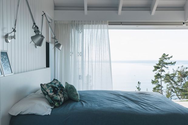 A serene coastal bedroom with a low bed dressed in blue-grey bedding, patterned and textured cushions, white vertical wall panelling, exposed white ceiling beams, and articulated wall-mounted reading lights. Sheer curtains frame expansive glazing with views toward the ocean and trees.