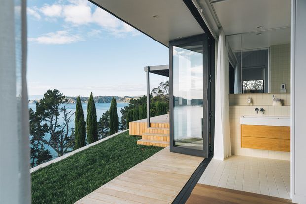 A contemporary waterfront-facing interior-exterior transition with large black-framed glazing and an open glass door leading onto a light timber deck and grassed terrace. The view includes water, trees, and a distant shoreline. At right is a compact bathroom with pale tiled walls and floor, a floating timber vanity, white basin, wall mirror, and exposed plumbing fixtures.