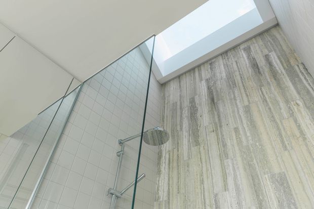Contemporary bathroom shower area viewed upward, featuring a frameless glass enclosure, wall-mounted rain shower, pale square wall tiles, a tall vertically textured stone-like feature wall, and a skylight providing natural light.