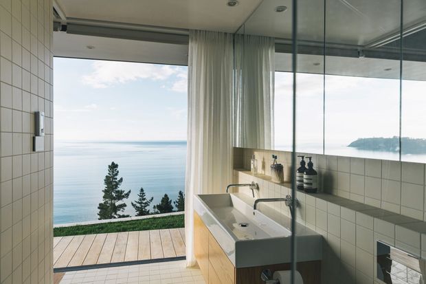 Contemporary bathroom with twin vessel basins, wall-mounted mirrored cabinetry, pale square wall tiles, and a glass-enclosed shower area. Full-height glazing and sheer curtains open to a timber deck with expansive ocean views, creating a bright, serene coastal setting.