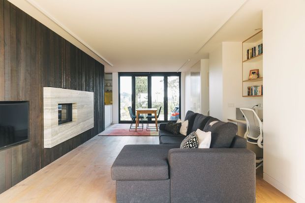 A contemporary open-plan living room with a large dark timber feature wall and integrated fireplace surround, light timber flooring, and a charcoal sectional sofa. The room extends toward a compact dining area and full-height glazed doors, with built-in open shelving and a small home office nook along the right wall.