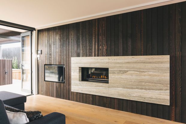 Contemporary living room featuring a dramatic dark timber-panelled feature wall, a wall-mounted television, and a linear fireplace set into a large pale stone surround. Warm timber flooring, neutral ceiling finishes, and extensive glazed doors opening to an outdoor area create a refined indoor-outdoor connection.