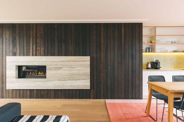 Open-plan living and dining area with a dramatic dark vertical timber feature wall, integrated linear fireplace in a pale stone surround, light timber flooring, and a dining nook beside open kitchen shelving. The room has warm concealed lighting, contemporary furnishings, and a strong contrast between dark wall panelling and light wood surfaces.