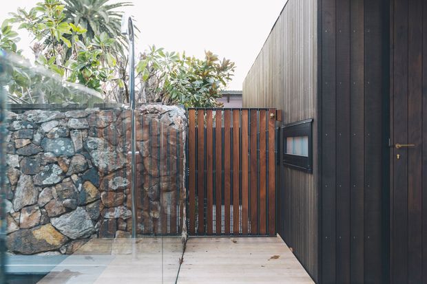 A narrow contemporary entrance passage with dark vertical timber cladding and a matching dark timber door on the right, a timber slatted gate ahead, and a natural rough-stone retaining wall on the left. Pale paved or timber-look flooring and lush planting create a refined, semi-enclosed approach.