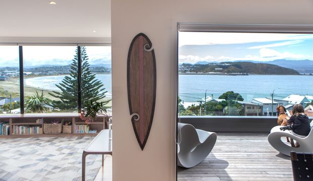 A bright coastal residential interior opening onto a timber deck with expansive water views. The image shows a living area with low built-in shelving, books and baskets, a patterned rug, contemporary sculptural lounge chairs, and a decorative surfboard mounted on a central wall. Large windows and sliding glazing connect the interior to the deck, where people are seated. The bay, shoreline and hills are visible beyond.