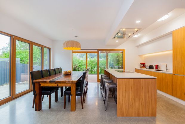 Bright open-plan kitchen and dining area with extensive timber-framed glazing, a long timber dining table, built-in bench seating, a large timber kitchen island with stools, and matching timber cabinetry. The space opens to a green outdoor area through glazed doors and is illuminated by recessed and pendant lighting.