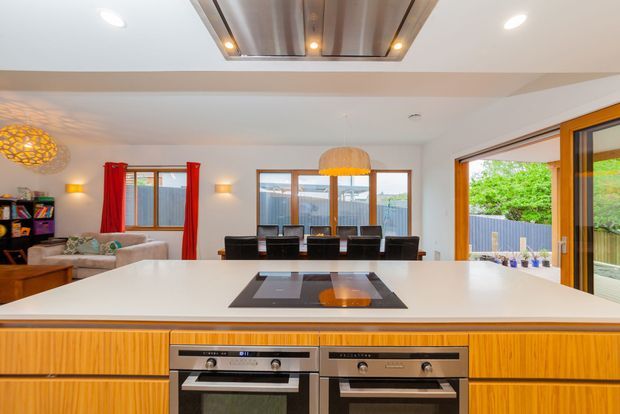 Bright open-plan kitchen and dining area viewed across a large island bench. The kitchen features warm timber-grain cabinetry, a white benchtop, integrated dark cooktop, and twin built-in stainless-steel ovens. Behind it is a dining table with pendant lighting, while a living area, red curtains, timber-framed glazing, and an open glazed door to greenery are visible. A decorative timber ceiling feature adds visual interest.