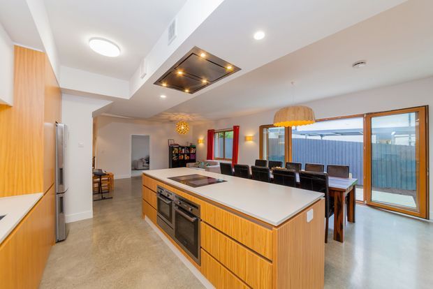 Bright open-plan kitchen and dining area with a large timber kitchen island, integrated appliances, pale benchtops, concrete-look flooring, and a long dining table beside full-height timber-framed glazing. A distinctive recessed ceiling feature with warm spotlights and pendant lighting adds visual interest.