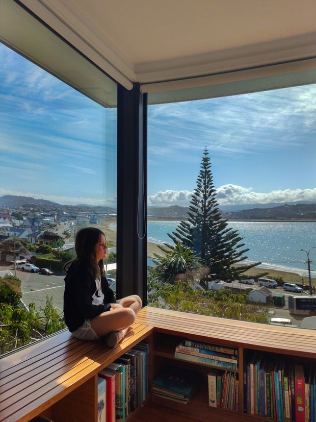 A bright interior corner with floor-to-ceiling corner glazing overlooking a coastal bay, hillside suburb and beach. A built-in timber window seat and bookcase occupy the foreground, with a seated person, roller blinds, and a tall evergreen tree visible outside. The space feels warm, airy and connected to the view.