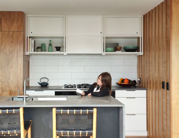 A contemporary kitchen viewed from the dining side, featuring a large grey stone island with integrated sink and seating, white flat-panel cabinetry, open shelving with ceramics, a white horizontal tile backsplash, timber wall panelling and tall timber cabinetry. Black fixtures and woven bar stools add contrast, while warm natural wood tones create a refined, inviting atmosphere.