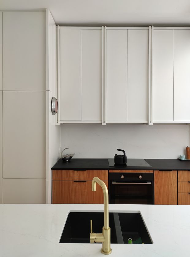 A bright, compact contemporary kitchen viewed across a large pale island benchtop. The island has a black undermount sink and brushed brass gooseneck tap. Along the rear wall are warm timber-grain lower cabinets, a black benchtop, integrated black cooktop and oven, and tall white upper cabinetry with slim framed doors and vertical trim details. The palette is restrained with white, light neutrals, black accents, warm timber, and brass hardware.