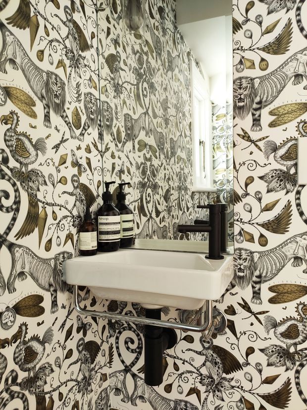 Close-up of a compact bathroom or powder room centered on a wall-mounted white ceramic basin with exposed chrome plumbing and a matte black tap. The walls are covered in highly patterned botanical and animal wallpaper in cream, black, grey, and muted olive tones. A narrow mirror reflects the basin area, while dark soap dispensers and a small window add functional detail. The space is eclectic, decorative, and visually bold.