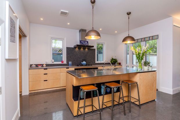 Bright contemporary kitchen with a large central island, integrated sink and dishwasher, timber-look cabinetry, dark stone benchtops and splashback, and three pendant lights over the island. The island includes seating for three, while glazed doors and windows bring in natural light. Polished dark flooring and white walls create a clean, refined appearance.