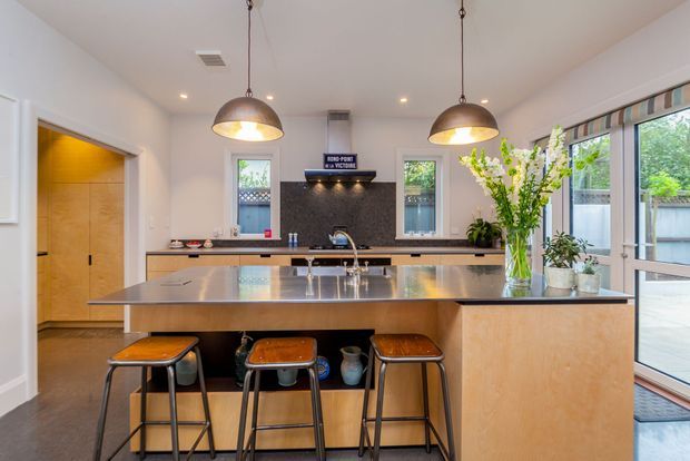Bright contemporary kitchen with a large central island, integrated sink and seating for three on simple stools. The room features light timber cabinetry, a dark textured backsplash, white walls, pendant lighting, and full-height glazed doors opening to an outdoor area. A pantry or utility space is visible through the doorway at left.