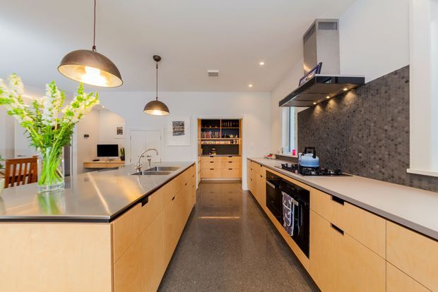 Bright contemporary kitchen with a large central island, extensive light timber cabinetry, dark polished flooring, and a dark textured tile splashback. The room includes integrated appliances, a gas cooktop with a stainless-steel rangehood, open shelving, pendant lighting, and a small dining area visible at the left.