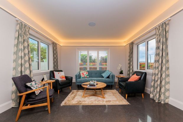 A bright, comfortably furnished living room with three large windows, patterned floor-length curtains, a central shuttered window, a teal sofa, two black leather armchairs, and a timber-framed lounge chair arranged around a round timber coffee table. Recessed perimeter ceiling lighting creates a warm glow, while the dark polished floor contrasts with light walls and a large neutral patterned rug.