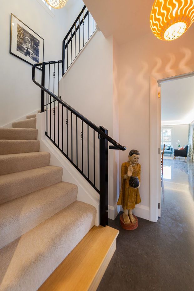 Bright interior view of a residential stairway and hallway. A carpeted staircase with timber stair edging rises beside a black metal balustrade and handrail, with white walls, framed artwork, and decorative pendant lights. A doorway opens toward a sunlit living room in the background, while a standing Buddha statue sits beneath the stairs.