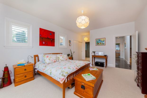 Bright bedroom with a traditional timber bed, matching wooden bedside furniture and chest, floral bedding, pale carpet, white walls, shuttered windows, framed artwork, and a decorative pendant light. A doorway leads to an adjoining hall or room.