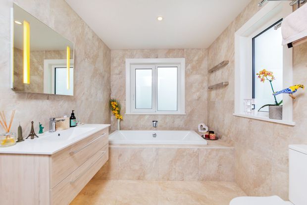 Bright, warmly finished bathroom with a large freestanding-style built-in bathtub beneath a central window, a wide floating timber-look vanity with double basins, illuminated wall mirror, tiled walls and floor, recessed ceiling lighting, and additional window shelving with decorative plants.