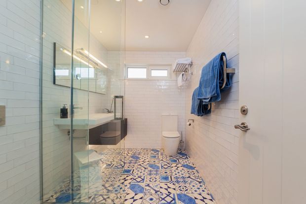 Bright contemporary bathroom with a walk-in glass shower, wall-mounted vanity, toilet, white subway-tile walls, and a bold patterned blue-and-white floor tile. Blue towels hang on wall-mounted rails, and a small high-level window provides natural light.