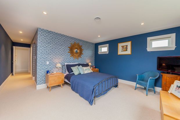 A spacious bedroom with deep blue painted walls, a patterned blue-and-white feature wall behind the bed, and soft beige carpet. The room includes a metal-framed bed with blue bedding, light timber bedside furniture, a sunburst mirror, framed artwork, a blue armchair, and a timber media console. Recessed ceiling lights and multiple small windows provide a bright, comfortable atmosphere.