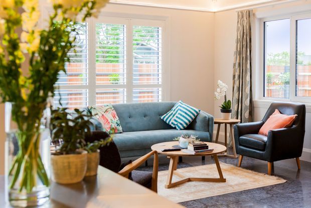 Bright, inviting living room with a blue-grey sofa, leather armchair, patterned cushions, timber coffee table, rug, indoor plants, and large windows with white plantation shutters. Neutral walls, decorative curtains, and classic ceiling trim create a polished contemporary interior with mid-century-inspired furnishings.
