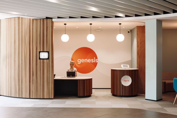 A contemporary commercial reception area with a curved black reception desk and matching cylindrical sign-in counter, both detailed with vertical timber slats. The space features pale stone or tile flooring, warm off-white walls with a large orange Genesis logo, globe pendant lights, a white structural column, and a slatted linear ceiling. A receptionist is seated behind the desk, and a small portion of lounge seating is visible at the right.